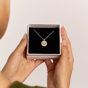 May include: A gold-colored pendant necklace with a circular charm featuring a detailed design, presented in a white box with a black interior. The necklace is displayed against a neutral background.