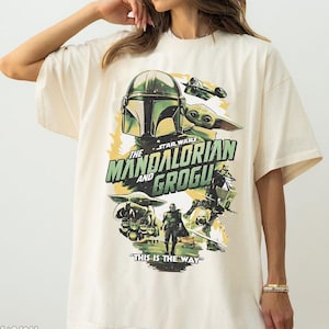 May include: Ivory-coloured t-shirt with a graphic from the Star Wars series, "The Mandalorian and Grogu." The design includes the Mandalorian's helmet, Grogu, and the text "This is the Way."