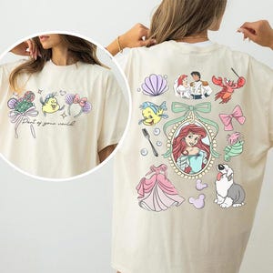 May include: Ivory-colored t-shirt featuring colorful cartoon graphics from a popular animated film. The front has a design with text, while the back showcases various characters and elements from the movie. The shirt is a comfortable, casual style.