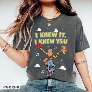 May include: A charcoal gray t-shirt featuring a graphic of a person with long blonde hair wearing a yellow plaid shirt and cow-print pants, holding a guitar and a red hat. The text reads I KNEW IT, I KNEW YOU, TS STORY 5, with cloud icons.