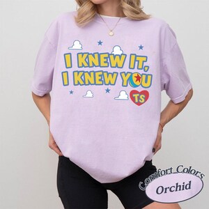 Op de afbeelding: Een lichtpaars t-shirt met korte mouwen voorzien van de gele tekst I KNEW IT, I KNEW YOU, een kleurrijke balafbeelding, een rood hart met de letters TS, en kleine blauwe sterren en witte wolken op de voorzijde van de borst.