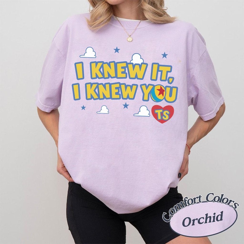 Op de afbeelding: Een lichtpaars t-shirt met korte mouwen voorzien van de gele tekst I KNEW IT, I KNEW YOU, een kleurrijke balafbeelding, een rood hart met de letters TS, en kleine blauwe sterren en witte wolken op de voorzijde van de borst.