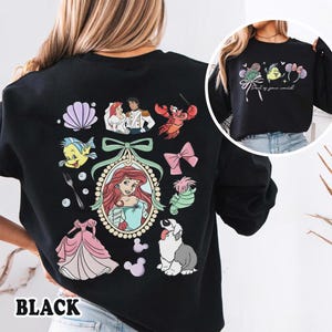 May include: Black sweatshirt featuring colorful illustrations of characters and elements from a popular animated film. The design includes a portrait of a princess, a seashell, a lobster, and other related graphics. The word "BLACK" is printed at the bottom.