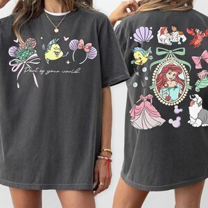 May include: Dark gray t-shirt featuring colorful illustrations from a popular animated film. The front displays a bow, a fish, and text that reads "Part of your world." The back has more characters and a portrait of a princess.