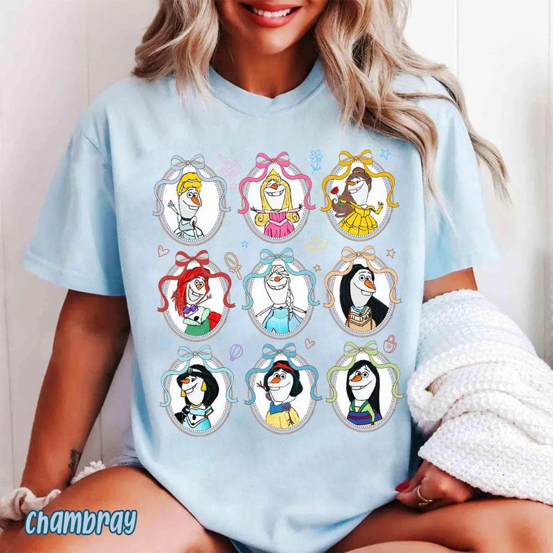 May include: Light blue t-shirt featuring cartoon illustrations of various characters in oval frames with bows. The illustrations include characters with blonde, red, and black hair, wearing dresses and accessories. The word "Chambray" is printed at the bottom.