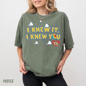 Op de afbeelding: Een olijfgroen t-shirt met korte mouwen en de gele tekst "I KNEW IT, I KNEW YOU", een kleurrijke balafbeelding, een rood hart met de gele letters "TS", en kleine witte wolkjes en blauwe sterren op de voorzijde van het kledingstuk.