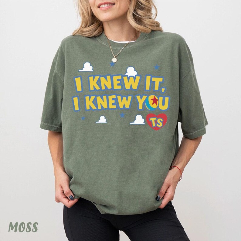 Op de afbeelding: Een olijfgroen t-shirt met korte mouwen en de gele tekst "I KNEW IT, I KNEW YOU", een kleurrijke balafbeelding, een rood hart met de gele letters "TS", en kleine witte wolkjes en blauwe sterren op de voorzijde van het kledingstuk.
