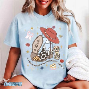 Peut inclure: Un t-shirt bleu clair avec une impression graphique repr&eacute;sentant une botte de cow-boy &agrave; motifs de vache, un chapeau de cow-boy rouge, un n&oelig;ud et des blocs avec le texte "TAYLOR" et "13". Le motif inclut des &eacute;toiles, des fleurs et des perles.