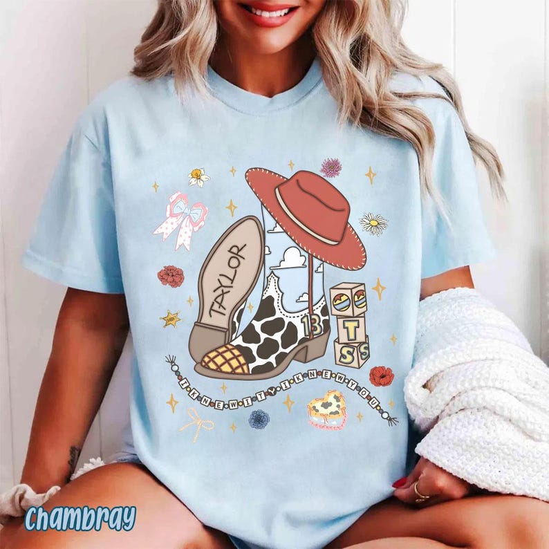 Peut inclure: Un t-shirt bleu clair avec une impression graphique repr&eacute;sentant une botte de cow-boy &agrave; motifs de vache, un chapeau de cow-boy rouge, un n&oelig;ud et des blocs avec le texte "TAYLOR" et "13". Le motif inclut des &eacute;toiles, des fleurs et des perles.