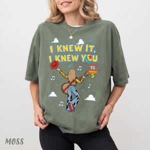 May include: An olive green t-shirt featuring a graphic of a person with long hair playing an acoustic guitar. The text reads I KNEW IT, I KNEW YOU and TS STORY 5. The design includes a red hat, musical notes, blue stars, and white clouds.