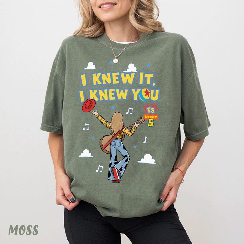 May include: An olive green t-shirt featuring a graphic of a person with long hair playing an acoustic guitar. The text reads I KNEW IT, I KNEW YOU and TS STORY 5. The design includes a red hat, musical notes, blue stars, and white clouds.