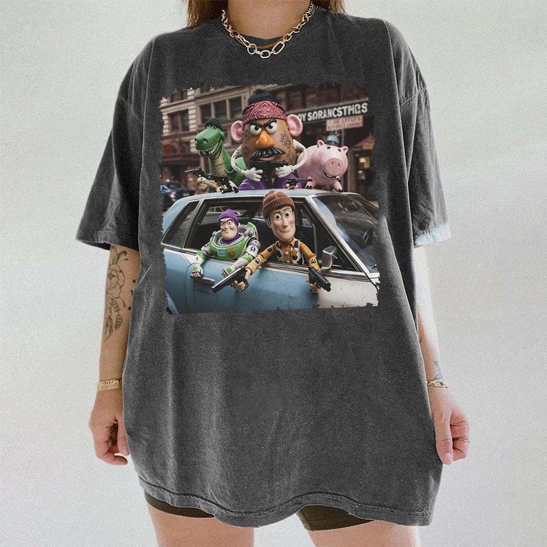 Op de afbeelding: Een antracietgrijs t-shirt met korte mouwen en een grafische print van Toy Story-personages, waaronder Mr. Potato Head, Rex, Hamm, Buzz Lightyear en Woody, afgebeeld in een sc&egrave;ne met een blauwe auto en de tekst SORANCSTHS.