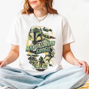 May include: White t-shirt with a Star Wars graphic featuring The Mandalorian and Grogu. The design includes the Mandalorian's helmet, Grogu, and the phrase "THIS IS THE WAY".
