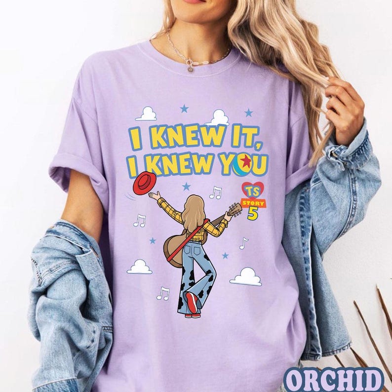 May include: A light purple t-shirt featuring a graphic of a person with long hair wearing a yellow plaid shirt and cow-print pants, holding a guitar and a red hat. The text reads I KNEW IT, I KNEW YOU, TS STORY 5, with clouds and musical notes.