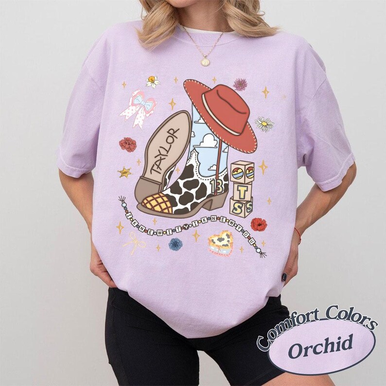 Peut inclure: Un t-shirt violet clair avec une impression graphique repr&eacute;sentant une botte de cow-boy &agrave; motifs de vache, un chapeau de cow-boy rouge, des blocs avec les lettres T, S et le chiffre 13, un n&oelig;ud, des fleurs et une perle avec le texte I KNEW YOU WERE TROUBLE.
