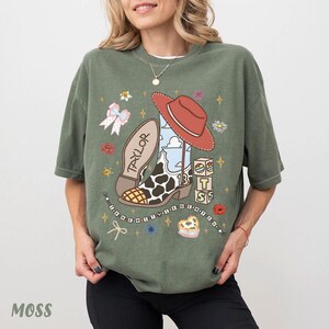 Peut inclure: Un t-shirt vert mousse avec une impression graphique repr&eacute;sentant une botte de cow-boy &agrave; motifs de vache, un chapeau de cow-boy rouge, un n&oelig;ud et des blocs avec les lettres T, S et le chiffre 13. Le texte TAYLOR et I KNEW YOU WERE TROUBLE est visible.