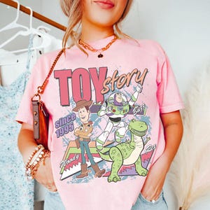 May include: Pink t-shirt featuring the words "Toy Story" in a retro font, with Woody, Buzz Lightyear, and Rex the dinosaur. The shirt also includes the text "Since 1995".