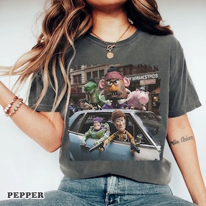 Op de afbeelding: Een antracietgrijs t-shirt met een grafische print van verschillende speelgoedfiguren in een auto. Het ontwerp toont een cowboy, een space ranger, een aardappelvormig personage, een groene dinosaurus en een roze varken met speelgoedwapens.