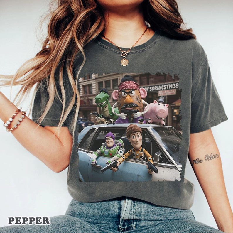 Op de afbeelding: Een antracietgrijs t-shirt met een grafische print van verschillende speelgoedfiguren in een auto. Het ontwerp toont een cowboy, een space ranger, een aardappelvormig personage, een groene dinosaurus en een roze varken met speelgoedwapens.