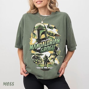 May include: Moss green t-shirt with a graphic design from The Mandalorian and Grogu. The design features the Mandalorian helmet, Grogu, and the text "The Mandalorian and Grogu" and "This is the Way".