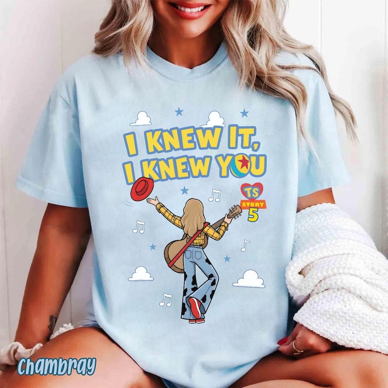 May include: A light blue t-shirt featuring a graphic of a person with long hair playing an acoustic guitar, wearing a yellow plaid shirt and cow-print bell-bottom pants. The text reads I KNEW IT, I KNEW YOU, TS STORY 5, with clouds and stars.