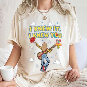 May include: A cream-colored t-shirt featuring a graphic of a person with long hair wearing a yellow plaid shirt and cow-print pants, holding a guitar and a red hat. The text reads I KNEW IT, I KNEW YOU, TS STORY 5, with clouds, stars, and music notes.