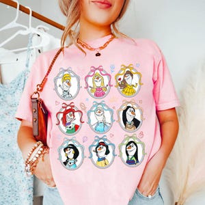 May include: Pink t-shirt featuring nine framed cartoon images of characters. Each image is a cartoon character with a bow and ribbon. The shirt is a light pink color and has a casual fit.