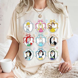 May include: Cream-colored t-shirt featuring a colorful graphic of cartoon characters in oval frames with bows. The design includes characters with various hairstyles and outfits. The shirt has short sleeves and a crew neck.
