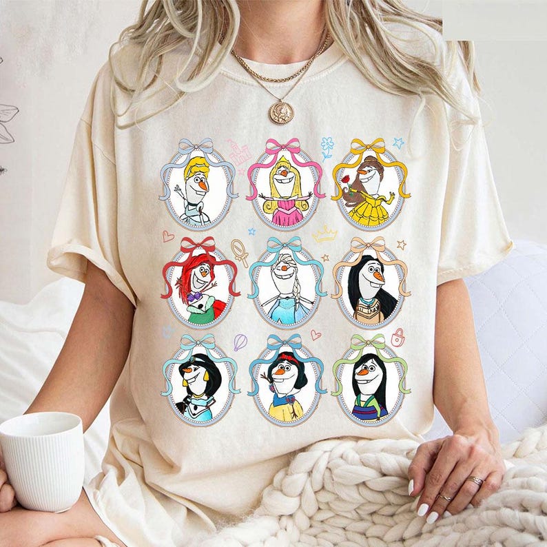 May include: Cream-colored t-shirt featuring a colorful graphic of cartoon characters in oval frames with bows. The design includes characters with various hairstyles and outfits. The shirt has short sleeves and a crew neck.