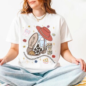 Peut inclure: Un t-shirt blanc avec une impression graphique repr&eacute;sentant une botte de cow-boy &agrave; motif de vache, un chapeau de cow-boy rouge et le nom TAYLOR. Le design inclut des blocs avec les lettres T, S, le nombre 13, des fleurs, des &eacute;toiles et des perles.