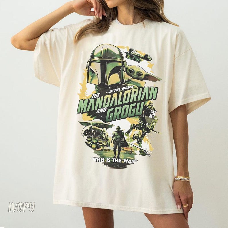 May include: Ivory-coloured t-shirt with a graphic from the Star Wars series, "The Mandalorian and Grogu." The design includes the Mandalorian's helmet, Grogu, and the text "This is the Way."