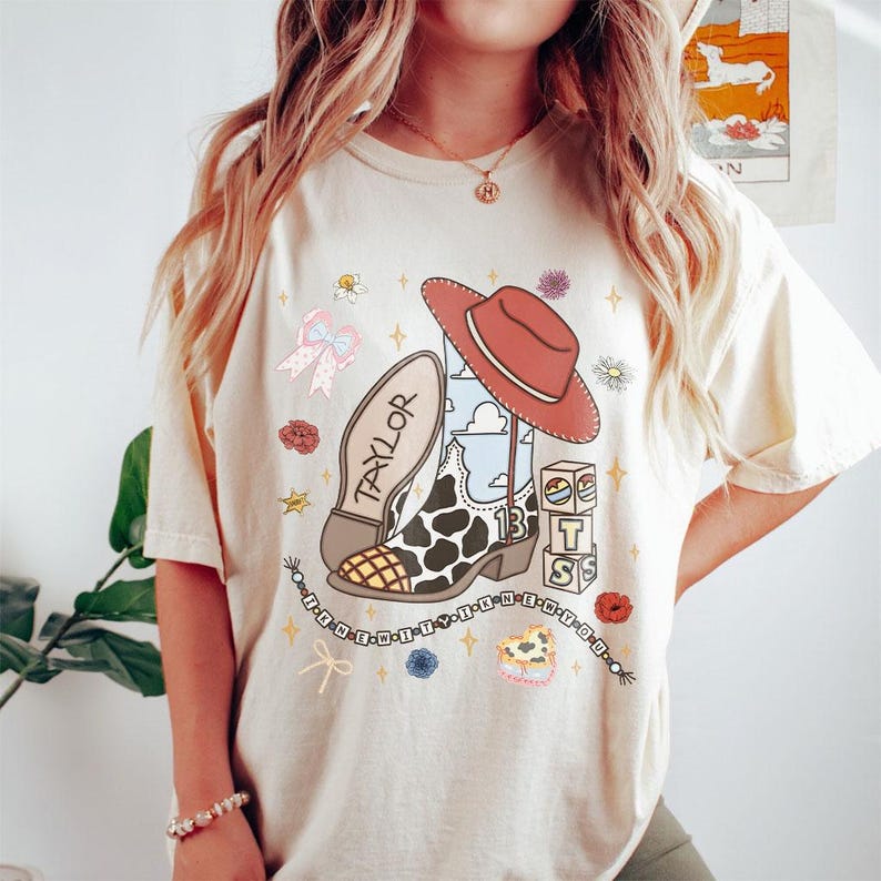 Peut inclure: Un t-shirt couleur cr&egrave;me avec une impression graphique repr&eacute;sentant une botte de cow-boy &agrave; motif de vache, un chapeau de cow-boy rouge, un n&oelig;ud et des blocs empil&eacute;s avec les lettres T, S et le chiffre 13. Le motif inclut le texte TAYLOR et des &eacute;toiles.