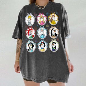 May include: Dark gray t-shirt featuring nine cartoon portraits of characters in oval frames with bows. The portraits include characters with red, blonde, and black hair, wearing crowns and dresses. The shirt has a relaxed fit.