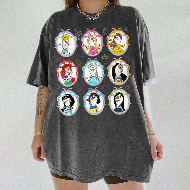 May include: Dark gray t-shirt featuring nine cartoon portraits of characters in oval frames with bows. The portraits include characters with red, blonde, and black hair, wearing crowns and dresses. The shirt has a relaxed fit.