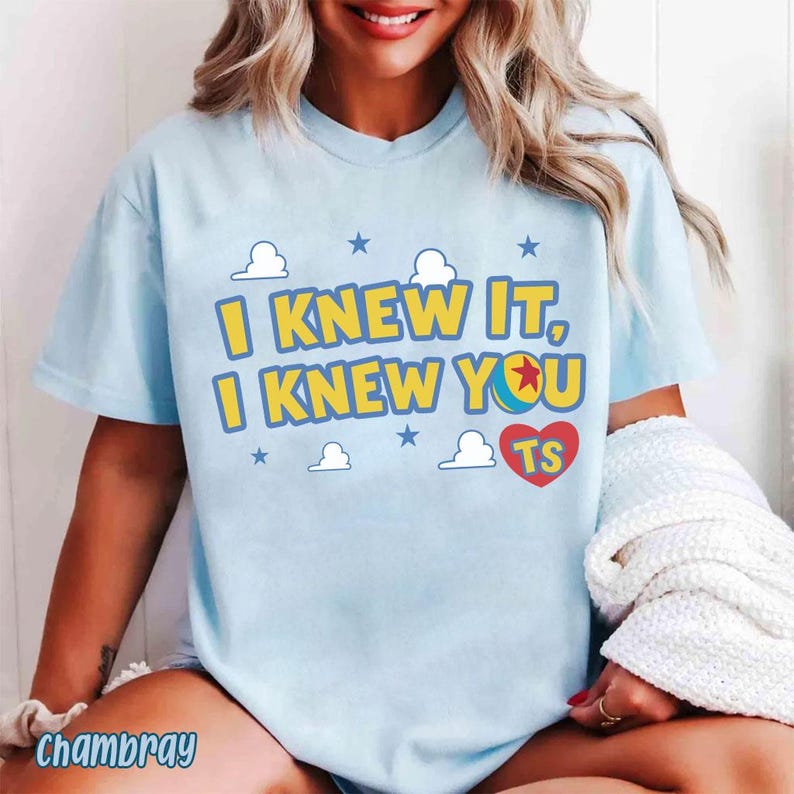 Op de afbeelding: Een lichtblauw t-shirt met de gele tekst I KNEW IT, I KNEW YOU, waarbij een kleurrijke balafbeelding de letter O vervangt. Het ontwerp bevat kleine witte wolken, blauwe sterren en een rood hart met de letters TS in het geel.