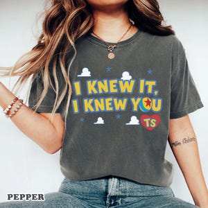 Op de afbeelding: Een antracietgrijs T-shirt met korte mouwen en de gele tekst I KNEW IT, I KNEW YOU, een kleurrijke balafbeelding die de letter O vervangt, een rood hart met de letters TS, en kleine witte wolken en blauwe sterren op de voorzijde gedrukt.