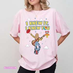 May include: A light pink t-shirt featuring a graphic of a person with blonde hair playing an acoustic guitar. The text reads I KNEW IT, I KNEW YOU, TS STORY 5. The design includes a red hat, blue cow-print pants, musical notes, and clouds.