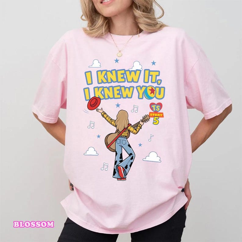 May include: A light pink t-shirt featuring a graphic of a person with blonde hair playing an acoustic guitar. The text reads I KNEW IT, I KNEW YOU, TS STORY 5. The design includes a red hat, blue cow-print pants, musical notes, and clouds.