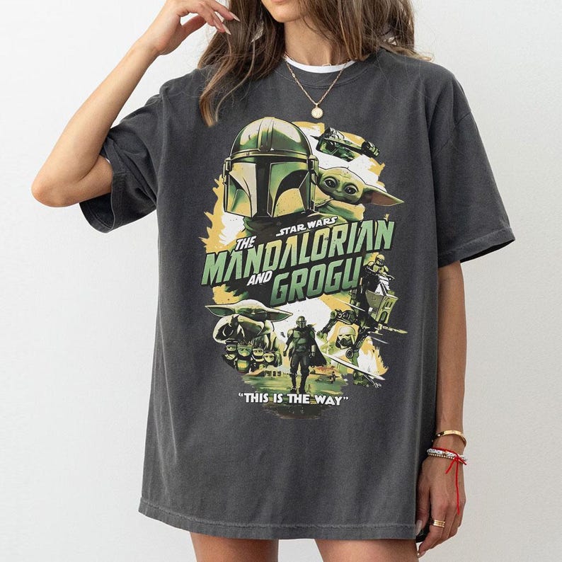 May include: Dark grey t-shirt with a graphic from the Star Wars series, "The Mandalorian and Grogu." The design includes the Mandalorian's helmet, Grogu, and the phrase "This is the Way."