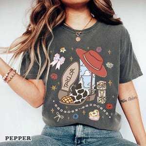 Peut inclure: Un t-shirt gris anthracite &agrave; manches courtes avec une impression graphique repr&eacute;sentant une botte de cow-boy &agrave; motifs de vache, un chapeau de cow-boy rouge, un n&oelig;ud rose et des cubes avec le chiffre 13. Le texte "TAYLOR" et "I KNEW IT I KNEW YOU" est imprim&eacute;.