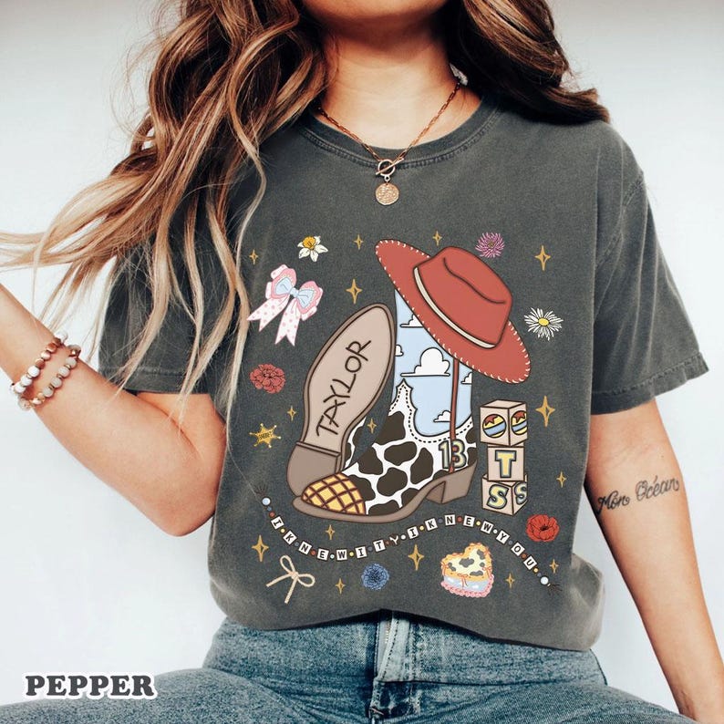 Peut inclure: Un t-shirt gris anthracite &agrave; manches courtes avec une impression graphique repr&eacute;sentant une botte de cow-boy &agrave; motifs de vache, un chapeau de cow-boy rouge, un n&oelig;ud rose et des cubes avec le chiffre 13. Le texte "TAYLOR" et "I KNEW IT I KNEW YOU" est imprim&eacute;.