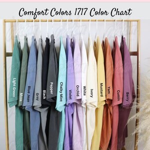 May include: A color chart displays a variety of Comfort Colors 1717 t-shirts in various hues. The shirts are arranged on hangers, showcasing colors like light green, blue jean, black, violet, and mustard. The text "Comfort Colors 1717 Color Chart" is at the top.