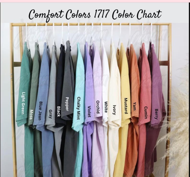 May include: A color chart displays a variety of Comfort Colors 1717 t-shirts in various hues. The shirts are arranged on hangers, showcasing colors like light green, blue jean, black, violet, and mustard. The text "Comfort Colors 1717 Color Chart" is at the top.