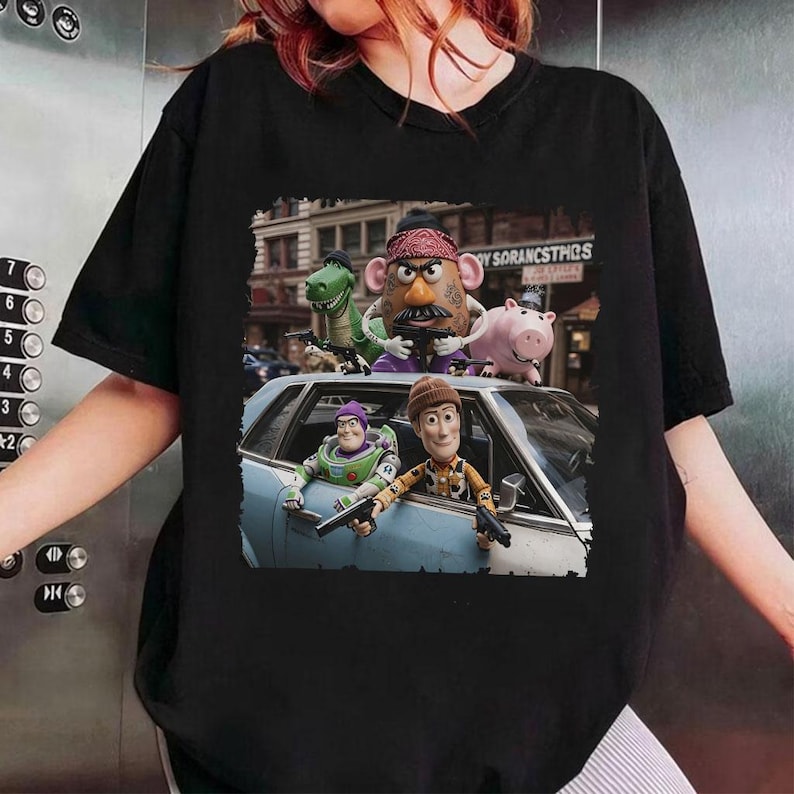 Op de afbeelding: Een zwart t-shirt met korte mouwen en een grafische print van vijf geanimeerde speelgoedfiguren uit Toy Story. De figuren zijn rond een lichtblauwe auto geplaatst, waarbij sommigen speelgoedpistolen vasthouden tegen een stadsachtergrond.