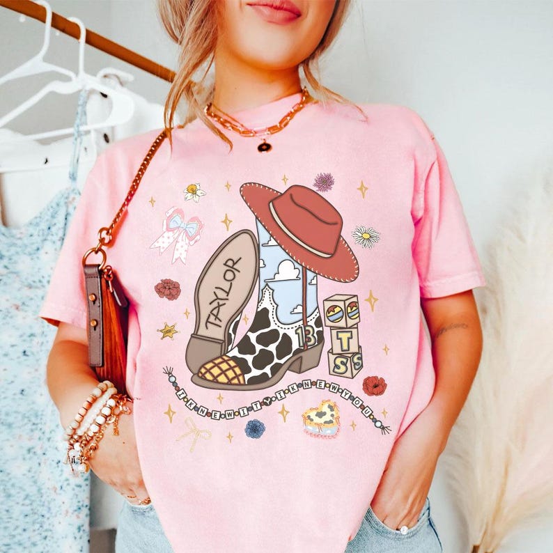 Peut inclure: Un t-shirt rose clair avec une impression graphique repr&eacute;sentant une botte de cow-boy &agrave; motif de vache, un chapeau rouge, des blocs avec les lettres T, S et un c&oelig;ur, le texte TAYLOR et I KNEW IT I KNEW YOU, entour&eacute;s de petites ic&ocirc;nes florales.
