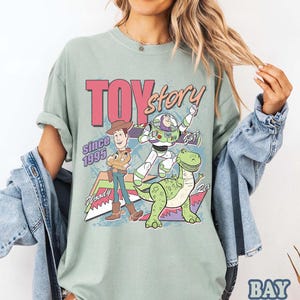 May include: A light green t-shirt featuring the words "TOY Story" in pink and the phrase "Since 1995". The shirt has a graphic of Woody, Buzz Lightyear, and Rex the dinosaur. The shirt is a comfortable, casual style.