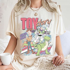 May include: Cream-colored t-shirt featuring a graphic of Toy Story characters. The design includes Woody, Buzz Lightyear, and Rex, with the words "Toy Story" and "Since 1995" in a retro style. The shirt has short sleeves.