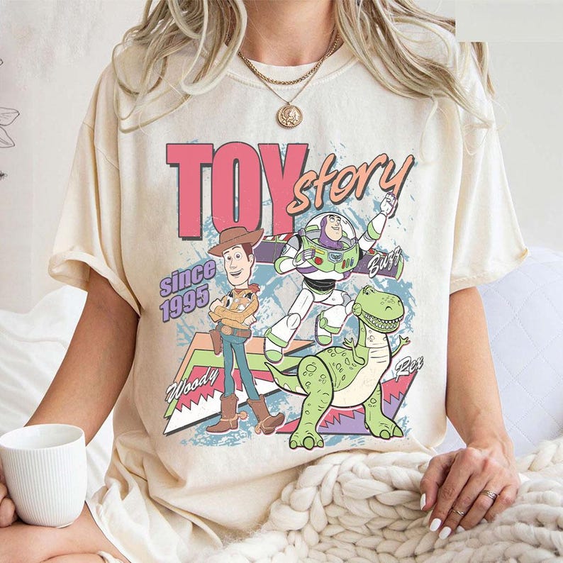 May include: Cream-colored t-shirt featuring a graphic of Toy Story characters. The design includes Woody, Buzz Lightyear, and Rex, with the words "Toy Story" and "Since 1995" in a retro style. The shirt has short sleeves.