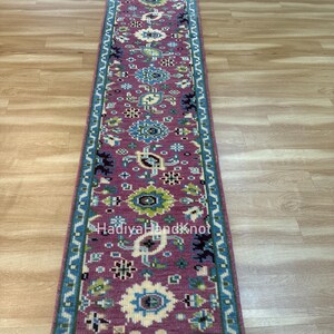 May include: A rectangular, hand-knotted runner rug with a detailed floral pattern. The rug features a deep rose-colored field with blue, green, and cream-colored floral motifs. The border is a teal blue with a repeating geometric design. The text "HadiyaHandKnot" is visible.