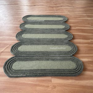 May include: A sage green and gray oval-shaped rug with a layered design. The rug is made of a soft, textured material and is placed on a wooden floor. The rug's design features multiple oval shapes stacked on top of each other.